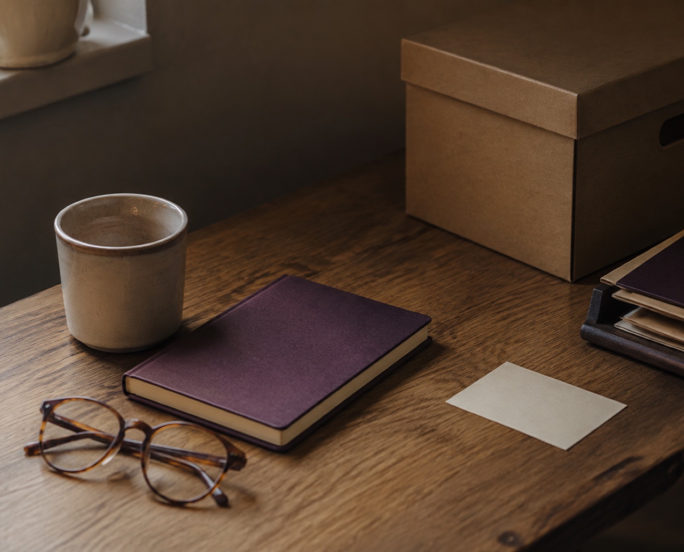 A closed plum notebook, reading glasses, a blank card and an archive box on a wooden desk.