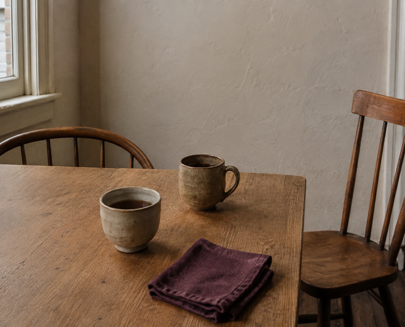 Two handmade cups and two wooden chairs at a modest kitchen table.
